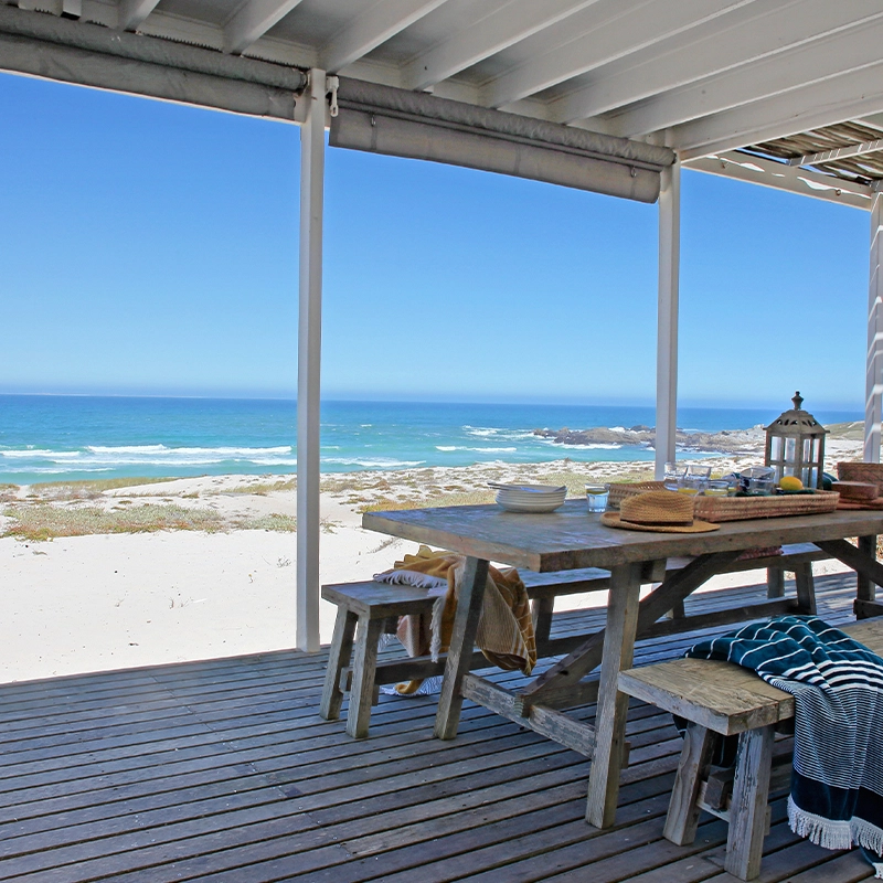 Perfect Hideaways — A rustic wooden deck with a long, weathered picnic table and benches overlooks a pristine white sand beach and turquoise ocean. A straw hat, striped towel, and lantern add relaxed charm under the shaded pergola, inviting slow seaside living. Yzerfontein, Not Forgotten.