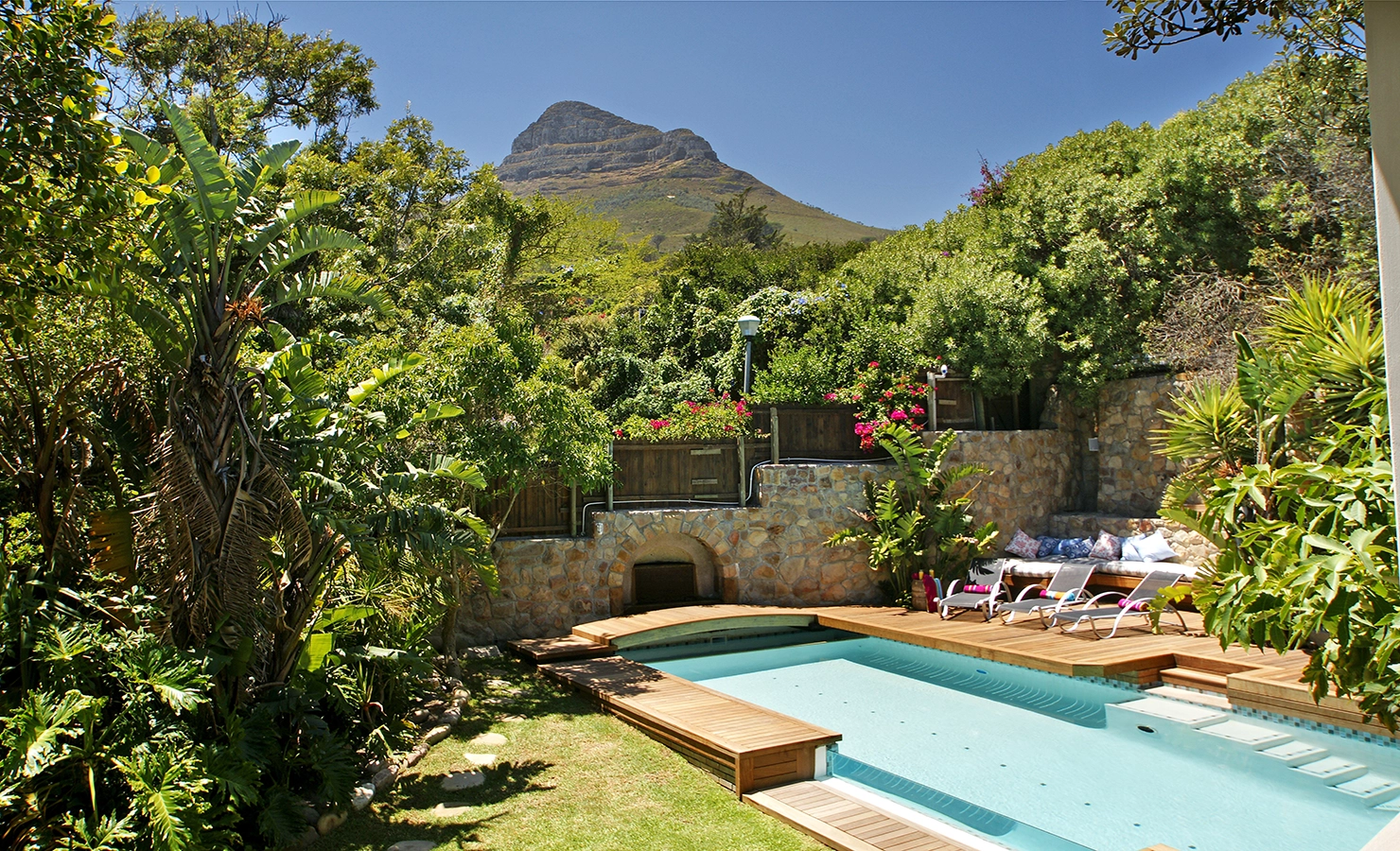 Perfect Hideaways: A serene poolside retreat surrounded by lush greenery, with a backdrop of Lion's Head mountain. The inviting wooden deck, sun loungers, and built-in seating create a tranquil space for relaxation in nature. Dolphin Cottage, Camps Bay.