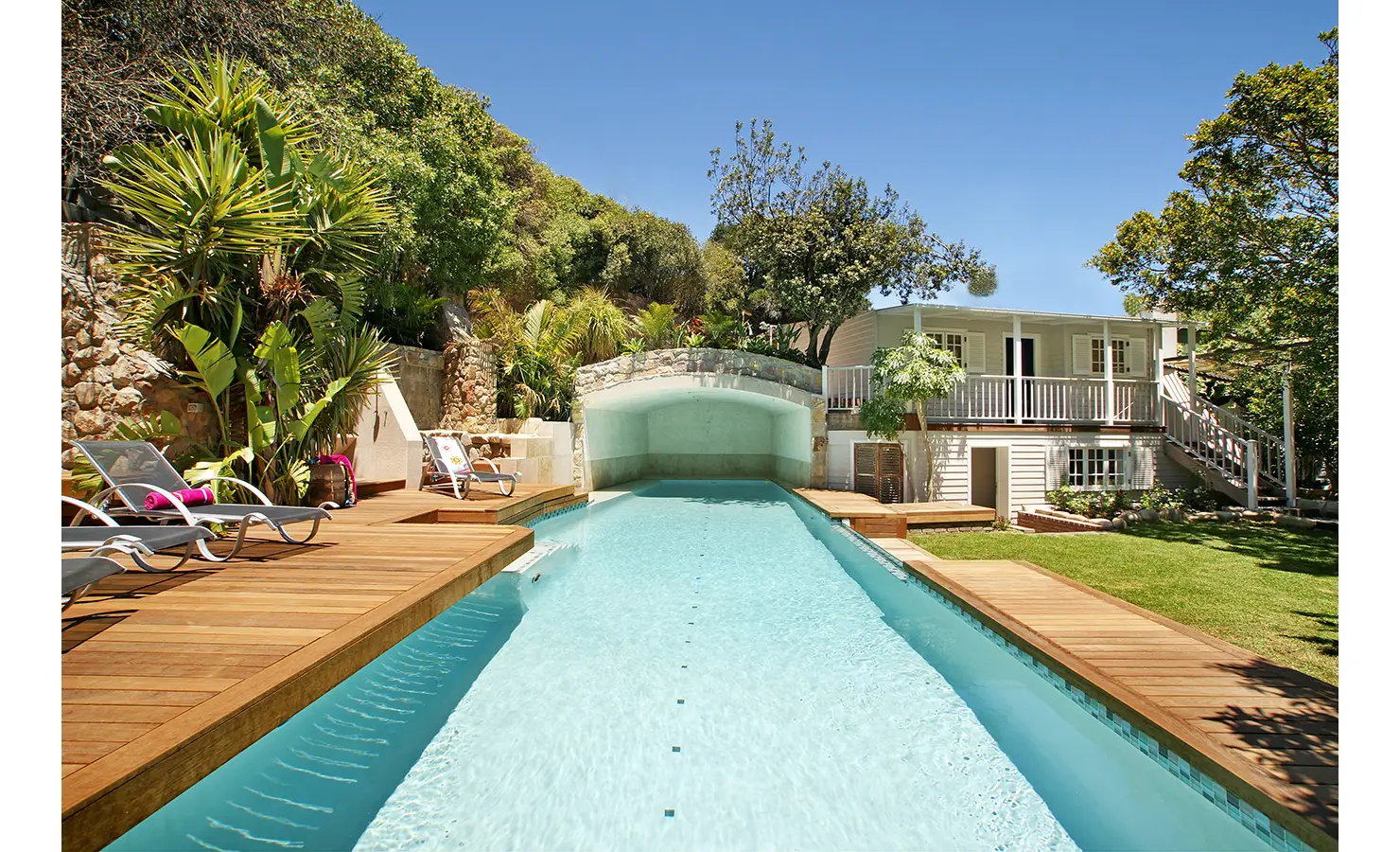 Perfect Hideaways: A refreshing swimming pool framed by wooden decking, lush tropical plants, and a charming stone arch, offering a peaceful escape under the Cape Town sun. Dolphin Cottage, Camps Bay.