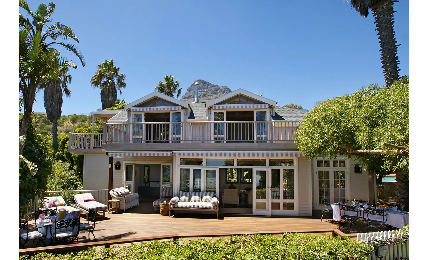 Perfect Hideaways: A stunning outdoor pool with a stone tunnel feature, surrounded by greenery and leading up to a classic Cape Cod-style beach house with wraparound balconies. Dolphin Cottage, Camps Bay.