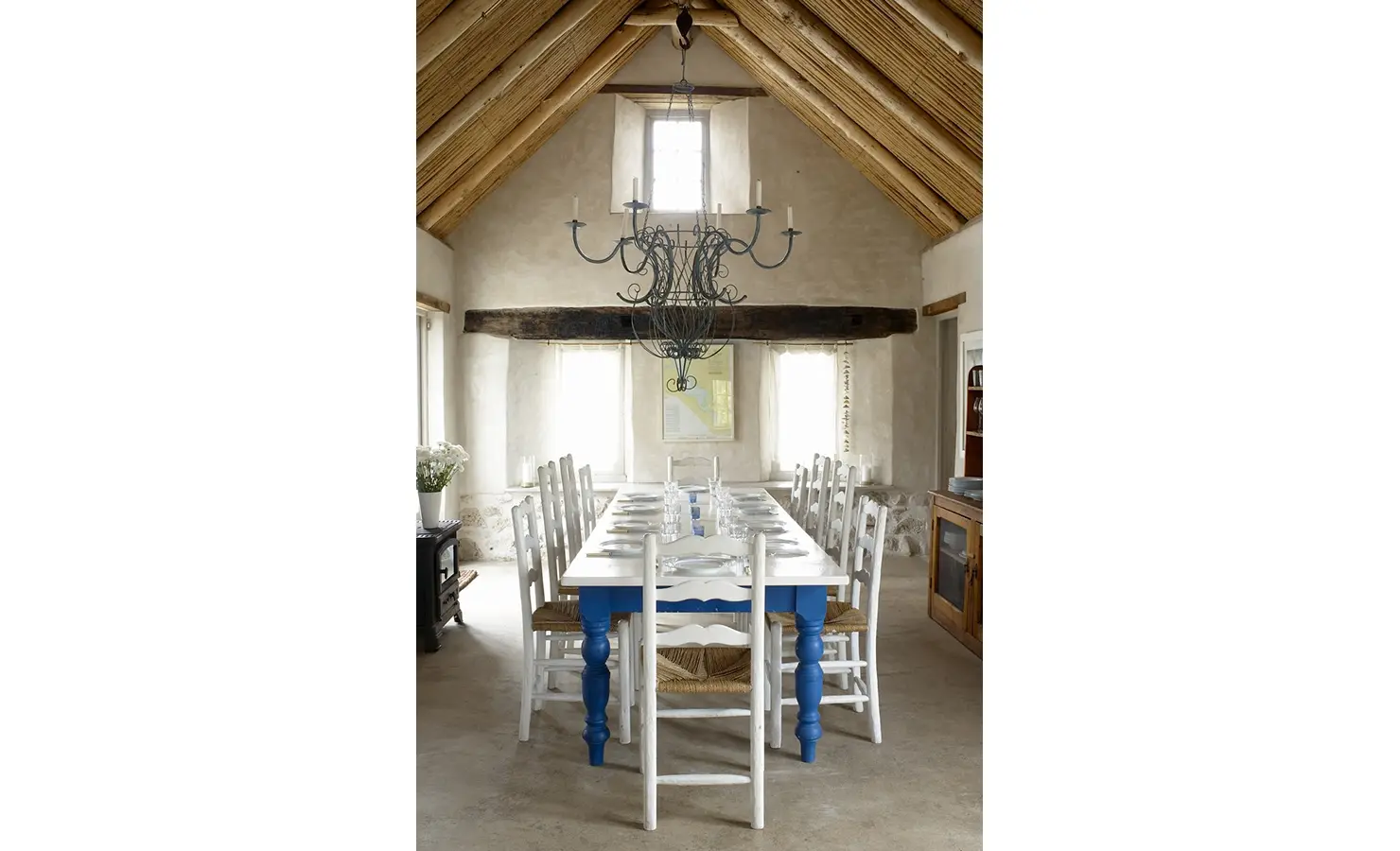 Perfect Hideaways: A beautifully rustic dining space with a vaulted thatch ceiling, exposed wooden beams, and a striking wrought-iron chandelier. A long wooden table with blue and white chairs is set for a welcoming meal in this airy, sunlit interior. Whalers Way, Churchhaven.