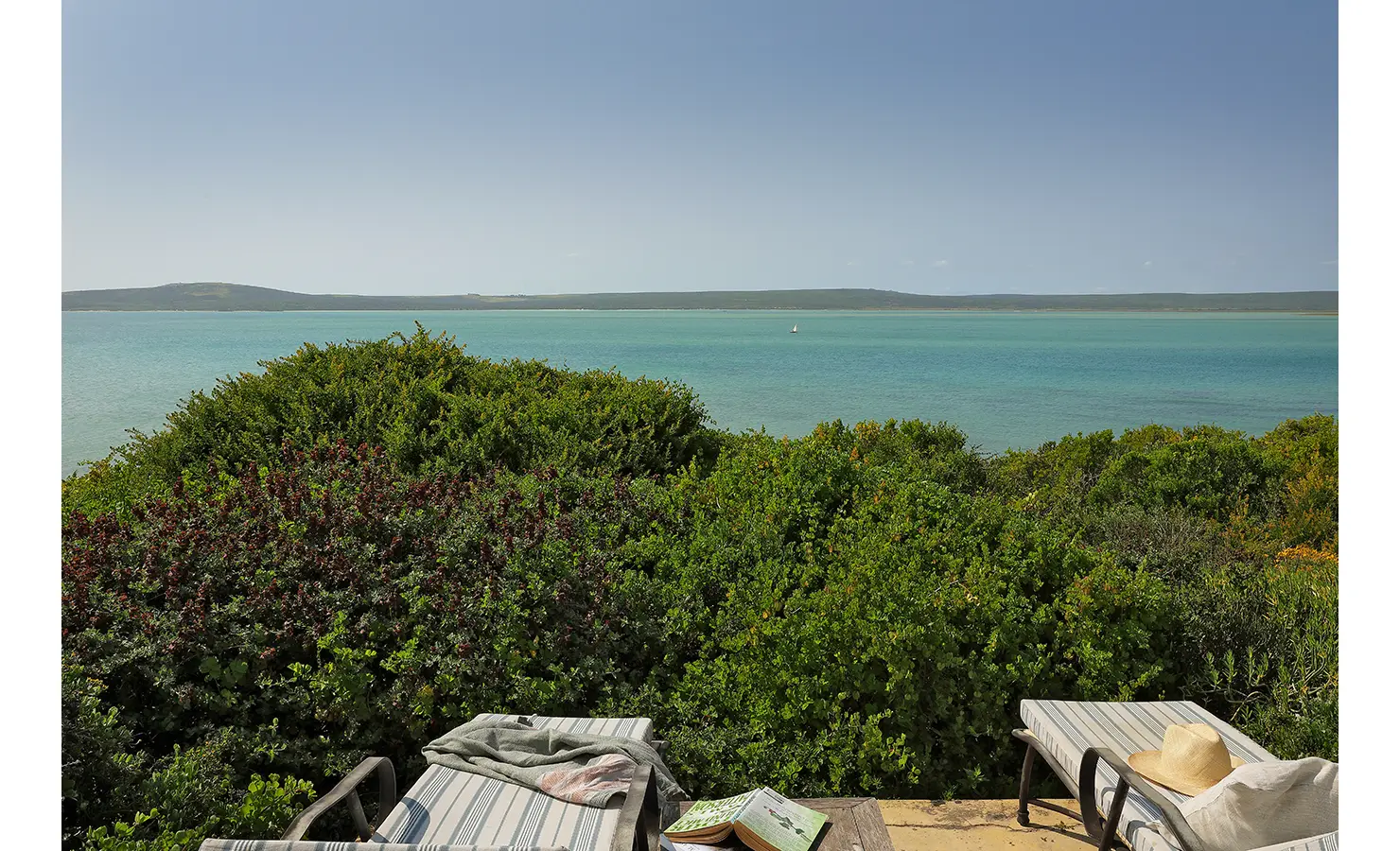 Perfect Hideaways: Two cushioned loungers rest beside dense coastal shrubs with an expansive view across the calm, glistening lagoon, a sailboat dotting the horizon in the distance. Churchhaven, Fynbos.