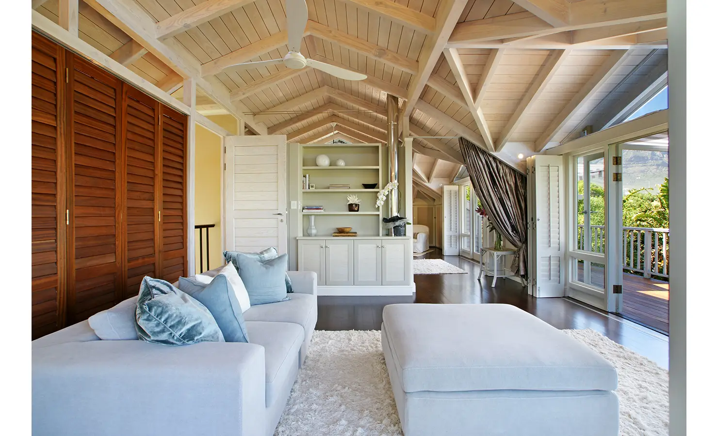 Perfect Hideaways: A tranquil loft-style lounge with soft neutral tones, plush seating, and exposed wooden beams, opening onto a private balcony with lush greenery. Dolphin Cottage, Camps Bay.