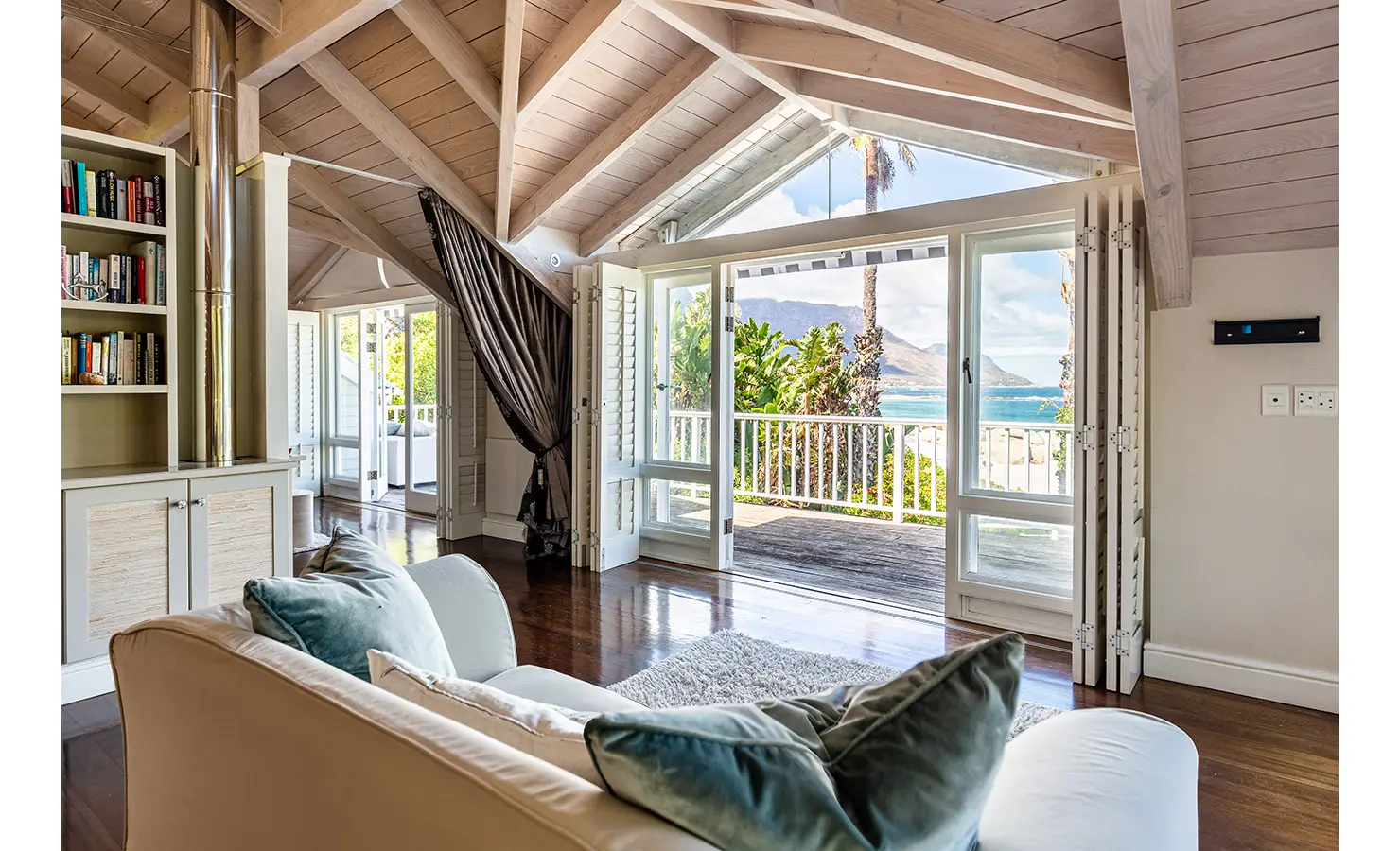 Perfect Hideaways: A light-filled coastal retreat with a high vaulted ceiling, folding doors leading to an ocean-view balcony, and soft blue and white décor creating a serene atmosphere. Dolphin Cottage, Camps Bay.