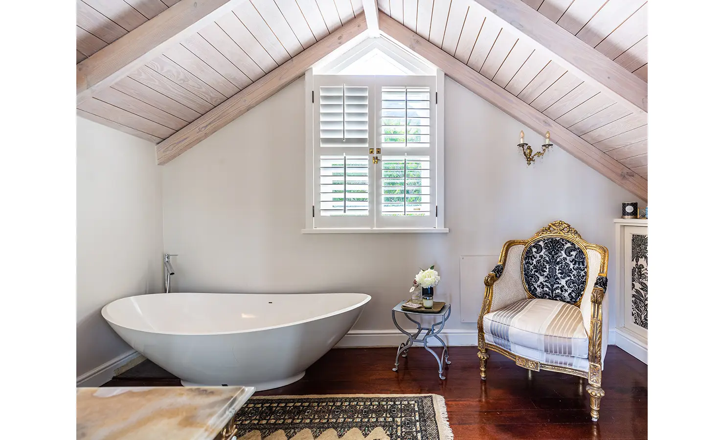 Perfect Hideaways: A sophisticated bathroom with a freestanding bathtub, golden-accented antique armchair, and sloped wooden ceiling, creating an elegant yet relaxed retreat. Dolphin Cottage, Camps Bay.