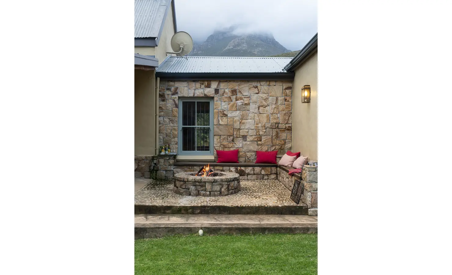 Perfect Hideaways: An intimate outdoor fire pit area surrounded by stone seating with red cushions, set against a rustic wall and misty mountains. Fizantakraal, Paarl.