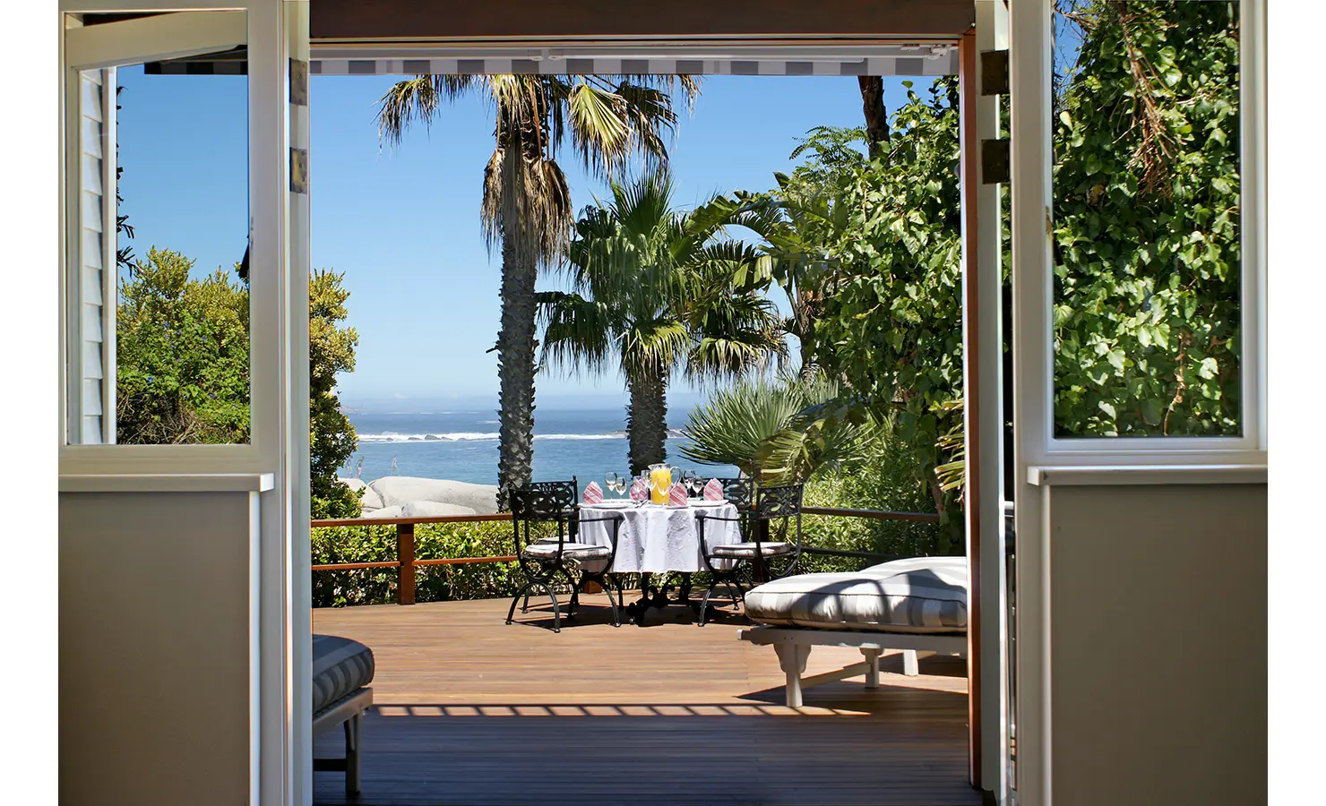 Perfect Hideaways: A sun-drenched deck with a private dining table, plush seating, and spectacular views of the ocean framed by swaying palm trees. Dolphin Cottage, Camps Bay.