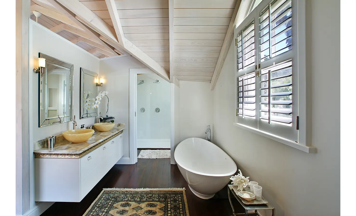 Perfect Hideaways: A refined en-suite bathroom with a modern freestanding bathtub, elegant double vanity with golden detailing, and a stylish attic window allowing in natural light. Dolphin Cottage, Camps Bay.