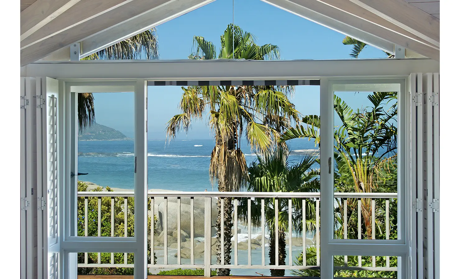 Perfect Hideaways: A picture-perfect ocean view framed by folding glass doors, with swaying palms and rolling waves creating an idyllic coastal escape. Dolphin Cottage, Camps Bay.