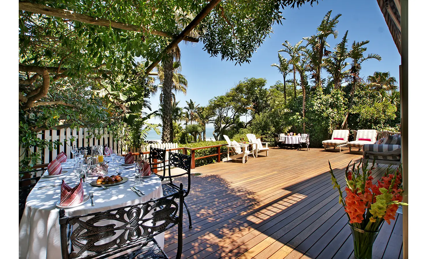 Perfect Hideaways: A spacious wooden deck shaded by lush greenery, featuring an elegant outdoor dining area, sun loungers, and cosy seating, all with a view of the ocean. Dolphin Cottage, Camps Bay.