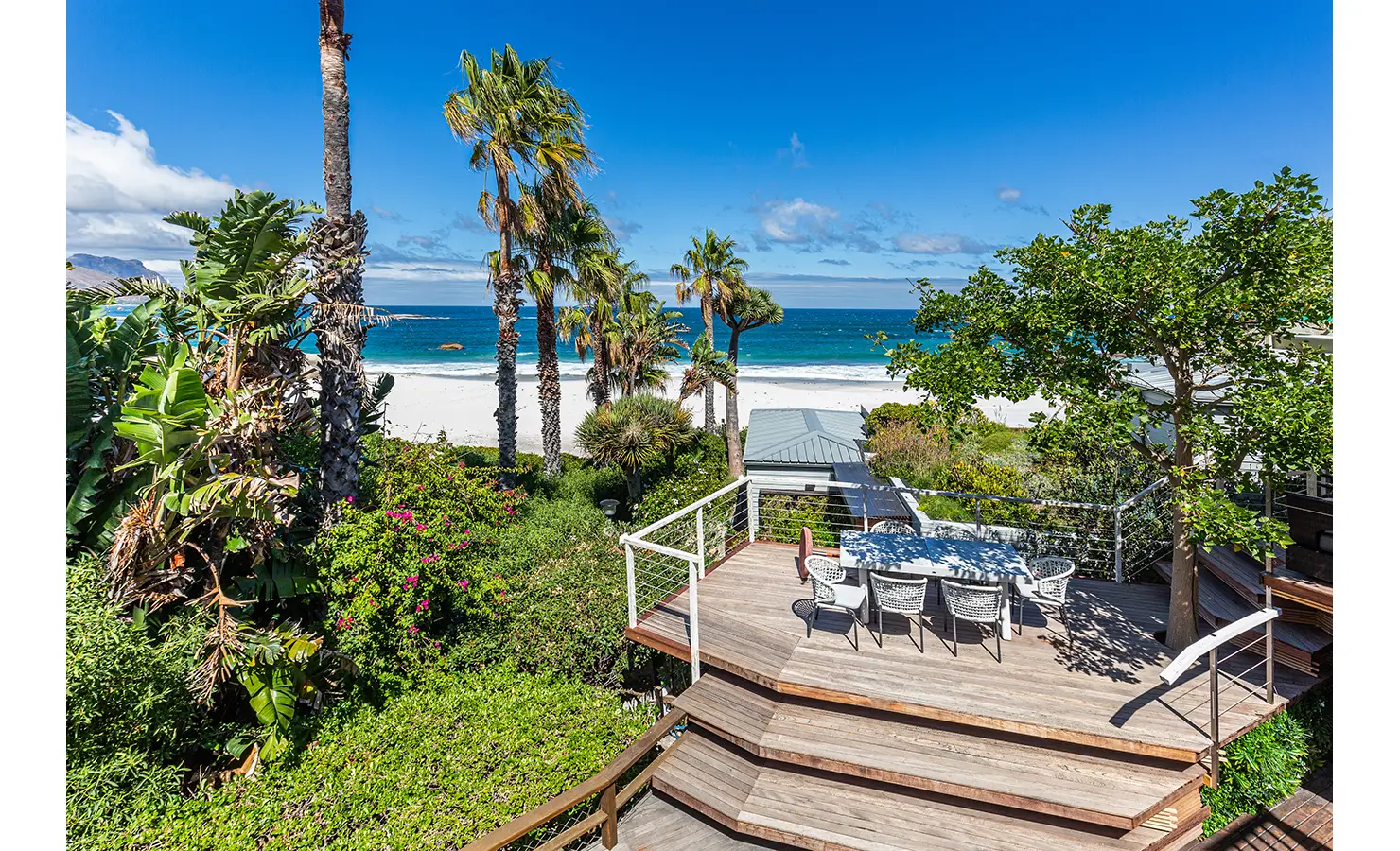 Perfect Hideaways: A breathtaking elevated terrace overlooking the white sands and turquoise waters of Camps Bay, surrounded by tropical palms and lush coastal vegetation. Dolphin Cottage, Camps Bay.