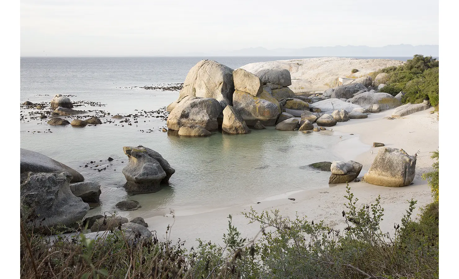 Perfect Hideaways: A secluded cove with turquoise waters and golden sand is framed by striking granite boulders, with penguins dotting the peaceful shoreline. Baxter House, Simons Town.