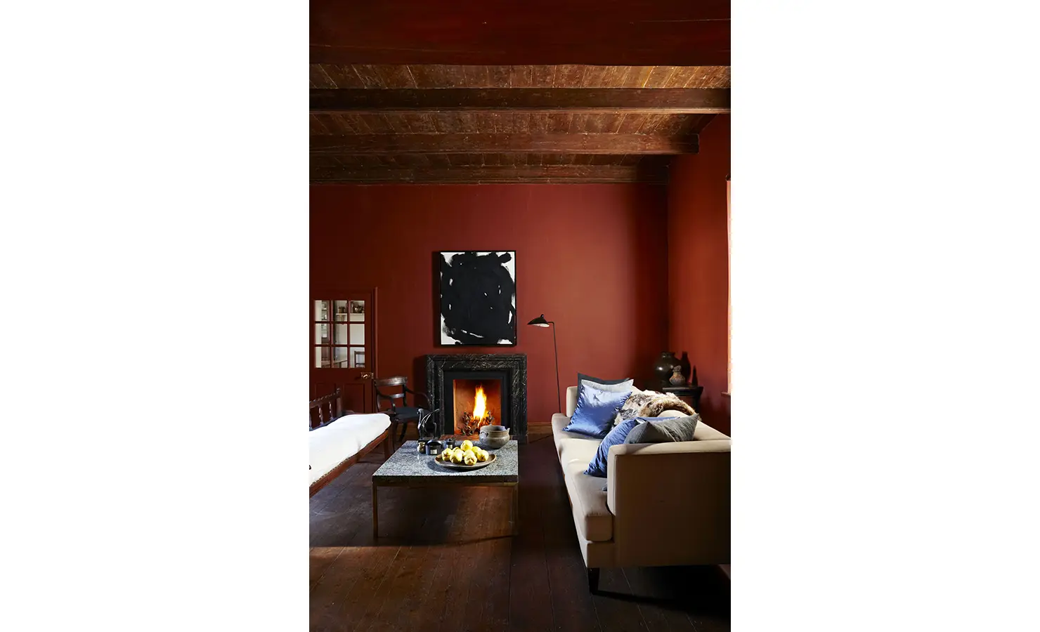 Perfect Hideaways: A striking living room with rich oxblood walls, wooden beams, a roaring fireplace, and plush seating adorned with blue and grey cushions, creating a dramatic yet cosy ambiance. Jonkmanshof, Montagu.
