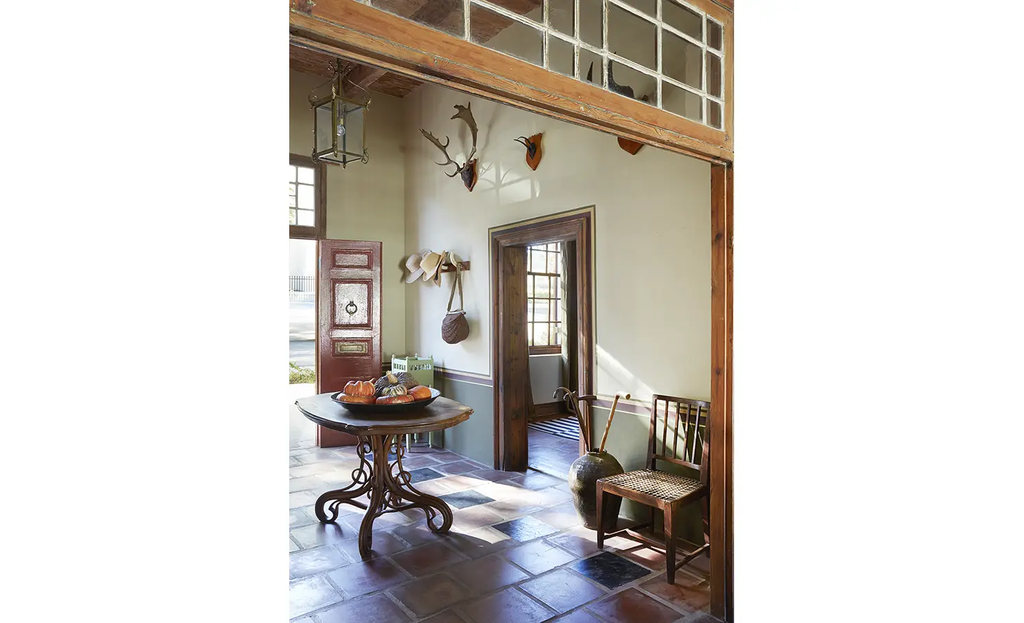 Perfect Hideaways: A light-filled entrance hall with a round wooden table topped with gourds, vintage chairs, antlers on the wall, and a glimpse of the red front door, welcoming and timeless. Jonkmanshof, Montagu.