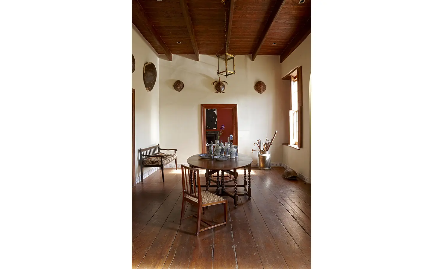 Perfect Hideaways: A serene dining space with a round wooden table, antique vases, woven chairs, and turtle shell décor along pale walls, framed by sunlight through sash windows. Jonkmanshof, Montagu.