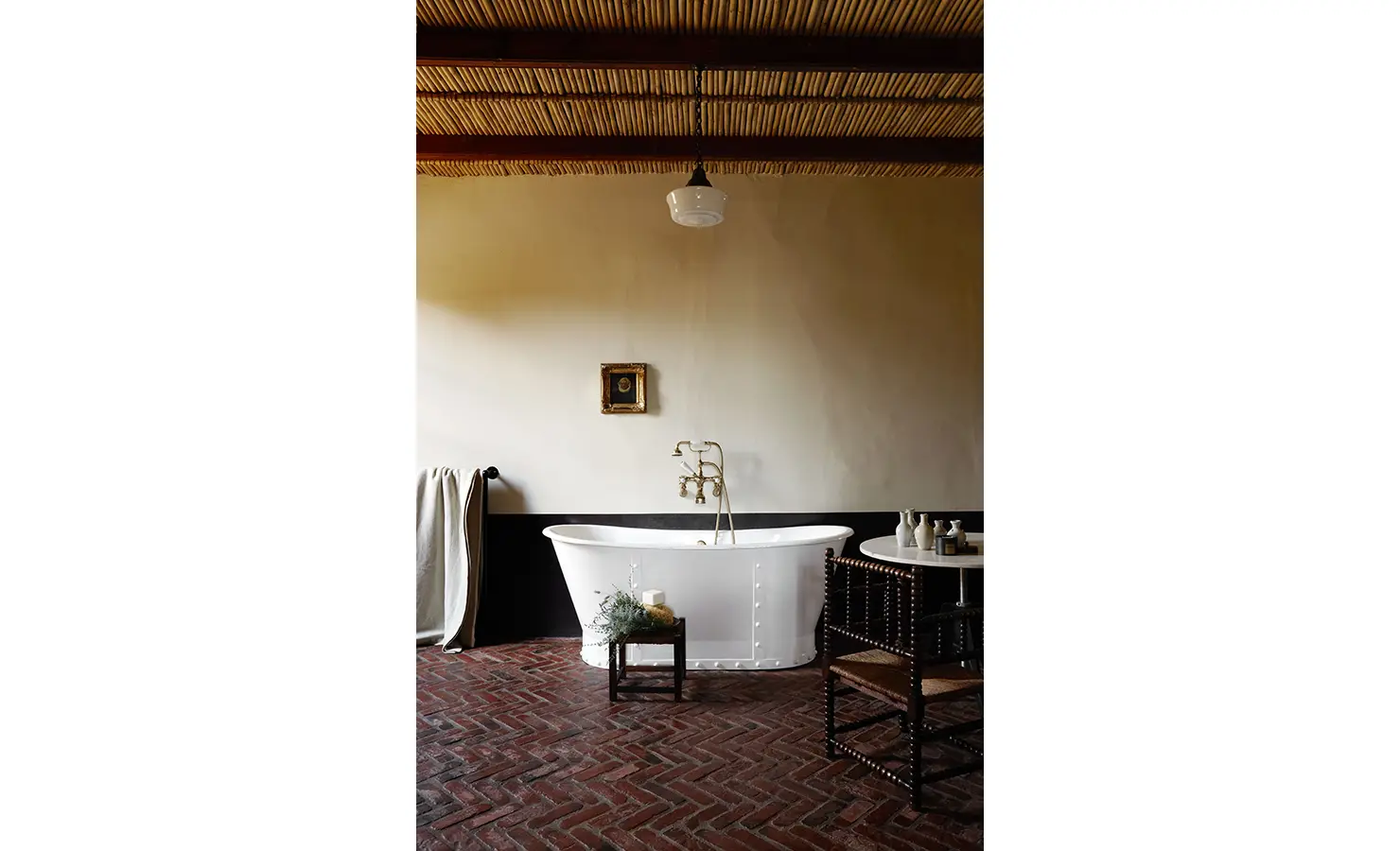 Perfect Hideaways: A dramatic bathroom with herringbone brick flooring, a freestanding white bathtub, woven reed ceiling, and a palette of earthy tones and textures. Jonkmanshof, Montagu.