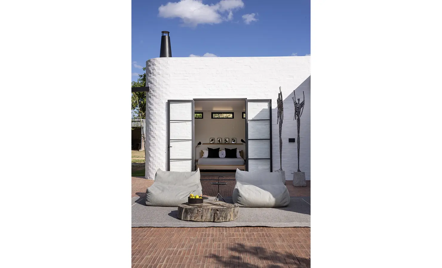Perfect Hideaways: A serene courtyard reveals double doors opening into a bedroom, flanked by tall sculptural artworks and minimalist beanbag loungers arranged around a rustic wooden table. STIL, Montagu.