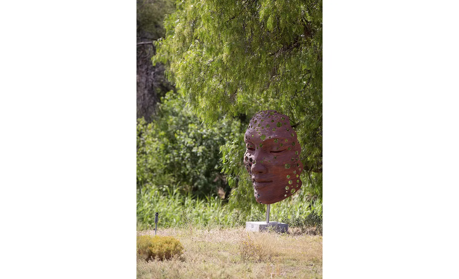 Perfect Hideaways: A large rust-toned sculptural face, punctuated with circular cut-outs, stands serenely under a tree in the natural garden, blending art and landscape. STIL, Montagu.