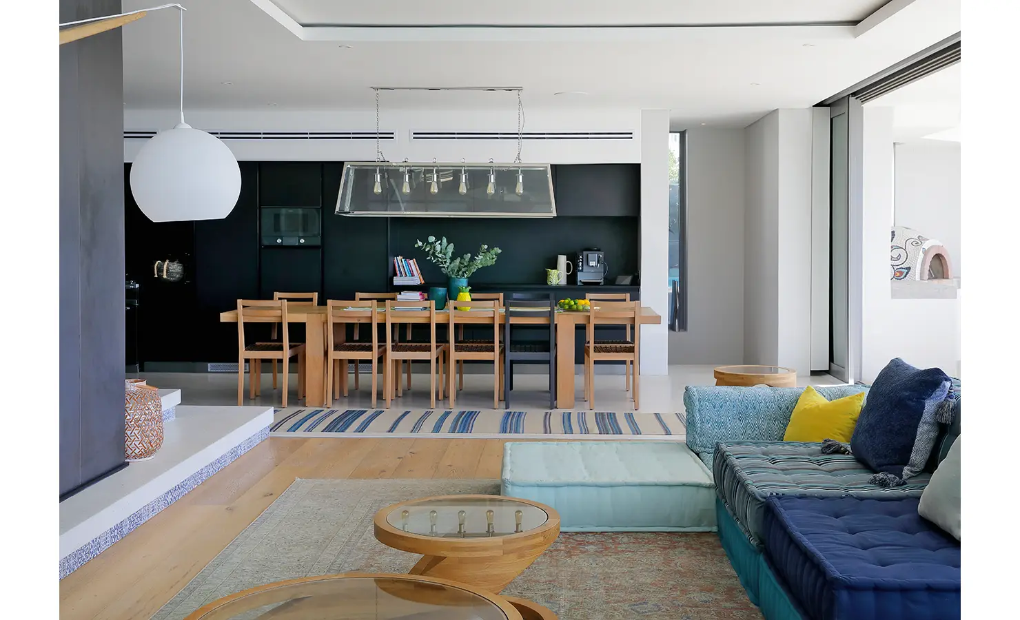 Perfect Hideaways: A stylish open-plan living and dining area with bold black cabinetry, a long wooden dining table, and colourful cushions on a modular sofa, creating a relaxed yet refined modern aesthetic. Bahari, Llandudno.