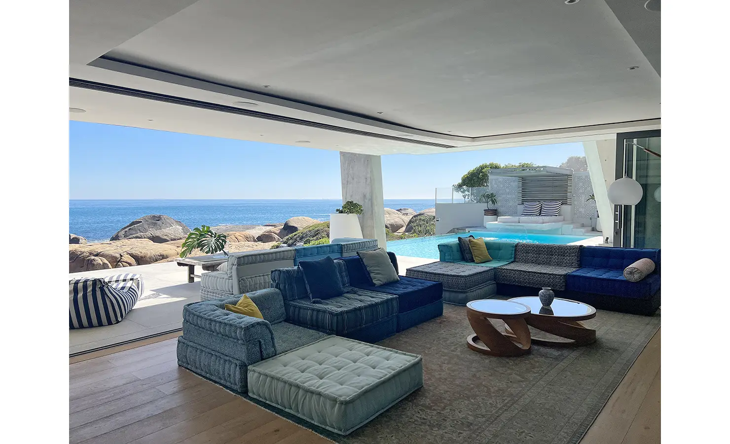 Perfect Hideaways: A breezy indoor-outdoor lounge with layered seating in textured blues and an infinity pool that flows seamlessly into dramatic ocean views and boulders beyond. Bahari, Llandudno.