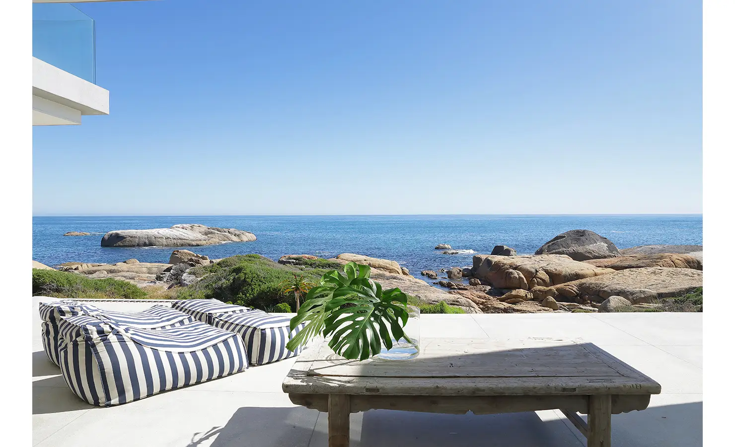 Perfect Hideaways: An alfresco dining table set against a panoramic seascape, with natural textures and striped seating blending into the rugged shoreline setting. Bahari, Llandudno.