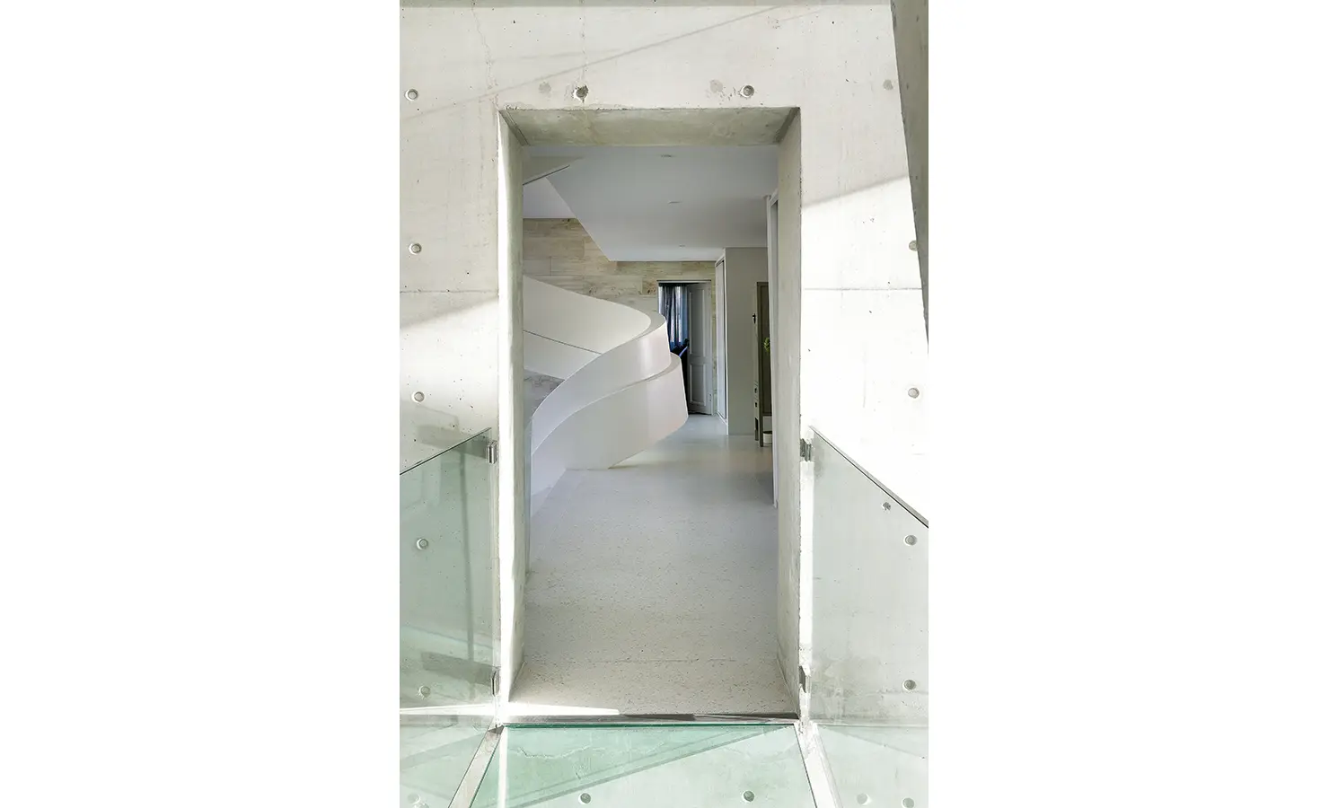 Perfect Hideaways: A modern concrete corridor with a glass floor and balustrades leads into the home, offering a glimpse of the sculptural spiral staircase beyond. Bahari, Llandudno.