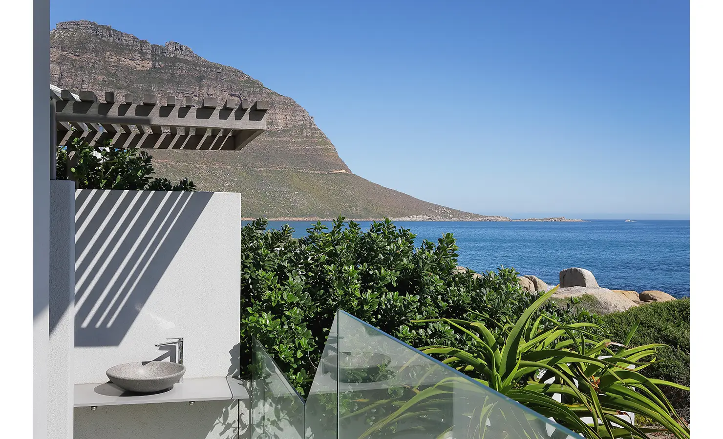 Perfect Hideaways: An outdoor stone basin set against a white wall with slatted shadows, framed by lush greenery and panoramic views of the Atlantic Ocean and dramatic mountain slopes. Bahari, Llandudno.