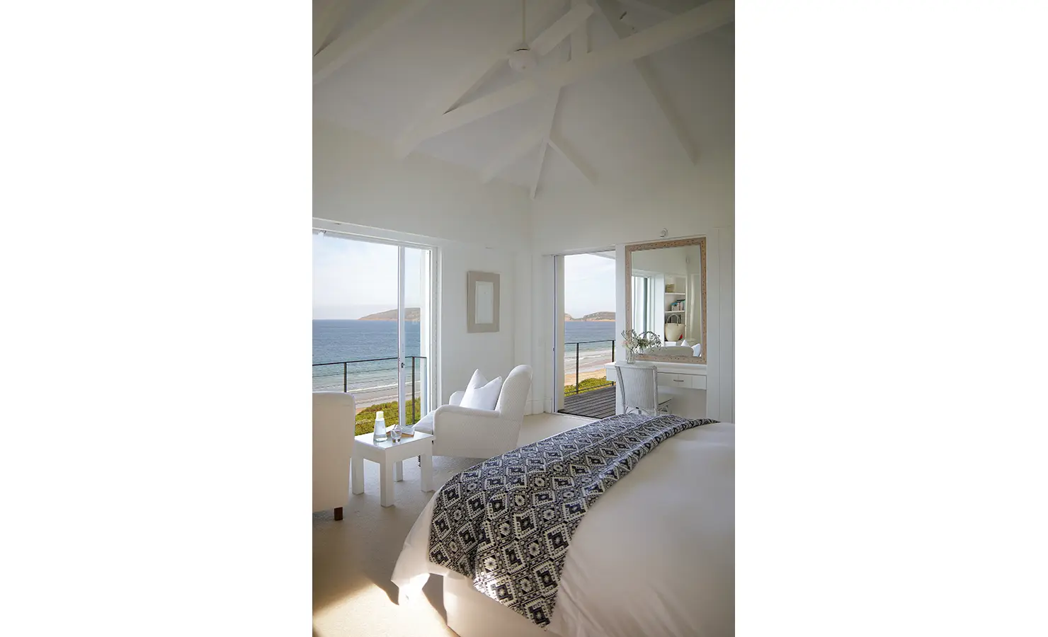 Perfect Hideaways: A bright bedroom featuring a cosy seating area, a mirrored dressing table, and stunning ocean views through expansive windows. Beachy Head House, Plettenberg Bay.