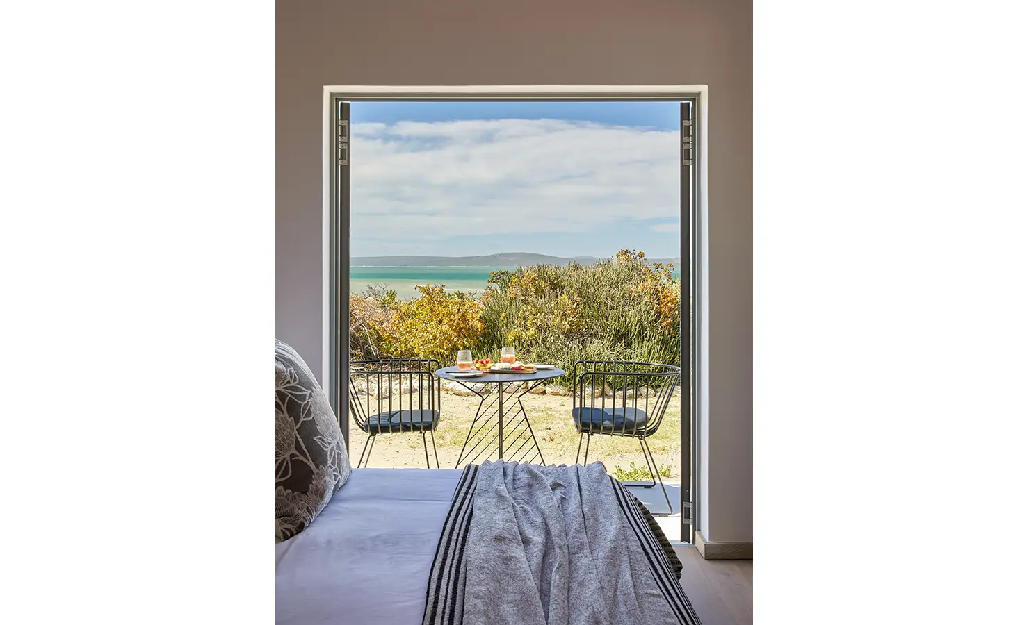 Perfect Hideaways: A close-up of an open bedroom door leading to a scenic patio with a small round table and two black wire chairs, set against a backdrop of lush greenery and the shimmering sea. Kalie's Cottage, Churchhaven.