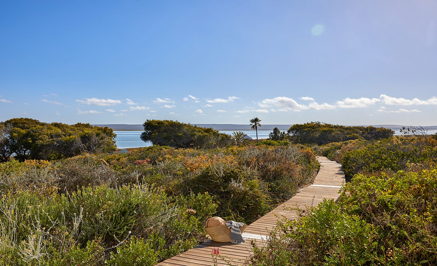Perfect Hideaways: A picturesque wooden boardwalk winding through the coastal fynbos, leading to the water’s edge. A woven beach bag and blanket rest on the path, inviting a tranquil seaside escape. Kalie's Cottage, Churchhaven.