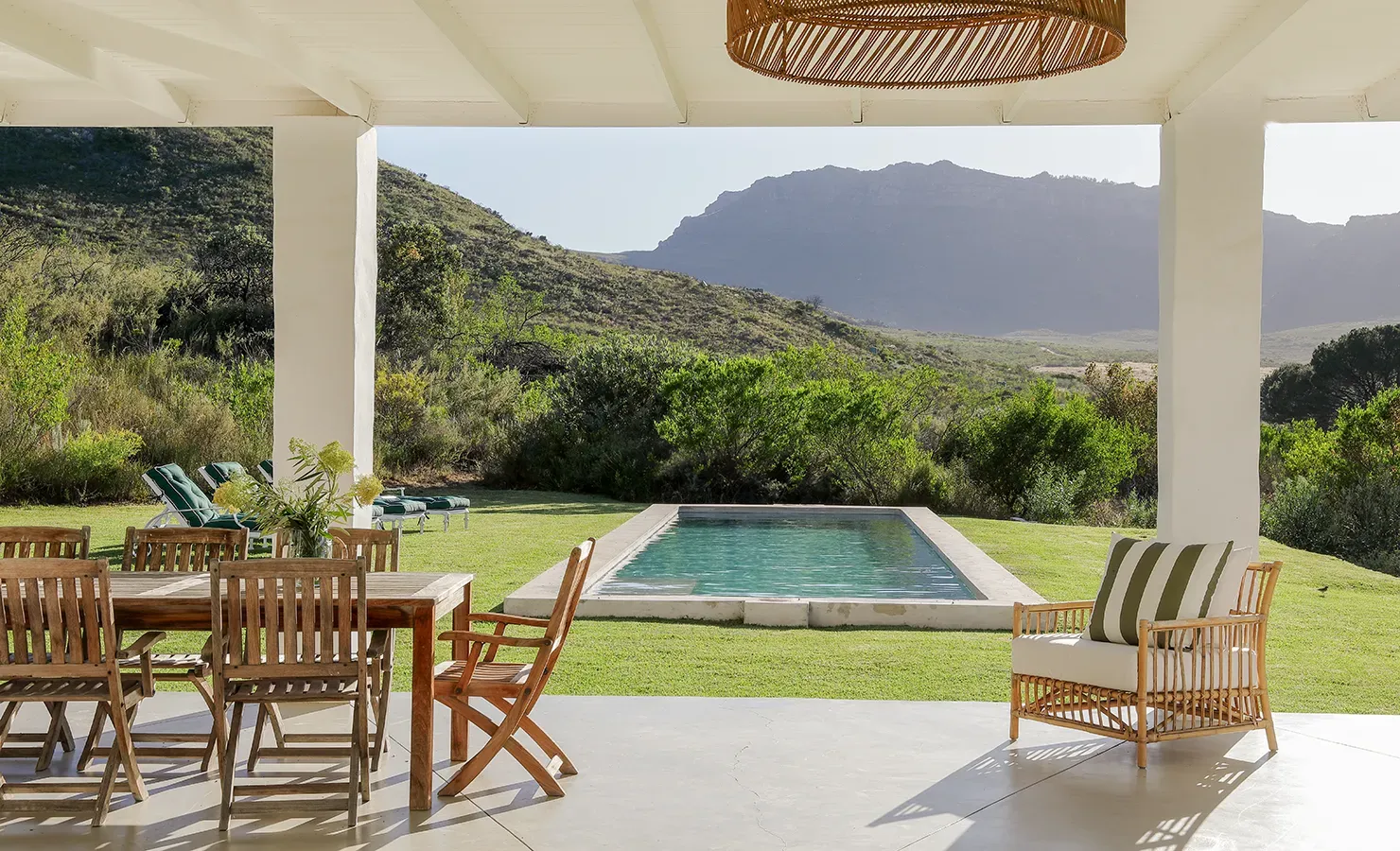 Buchubos Villa verandah with an outdoor wooden dining table and chairs overlooking the lap pool and green rolling lawn that is surrounded by the natural landscape and distant mountain range.