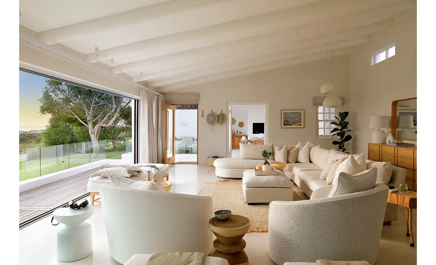 Perfect Hideaways: A bright and airy living room with a large L-shaped sofa and plush armchairs looks out through sliding doors onto a garden and tree-lined coastal view. Kinsail Beach Villa, Langebaan.