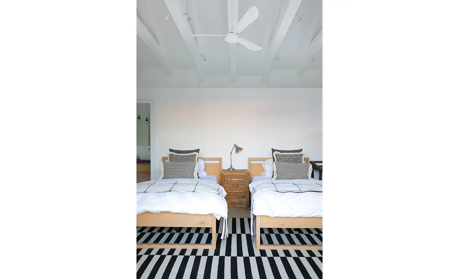 Perfect Hideaways: A minimalist bedroom with two twin beds, featuring wooden frames, patterned cushions, and a striped black-and-white rug. The room is illuminated by a ceiling fan and natural light. Ola Kala, Plettenberg Bay.