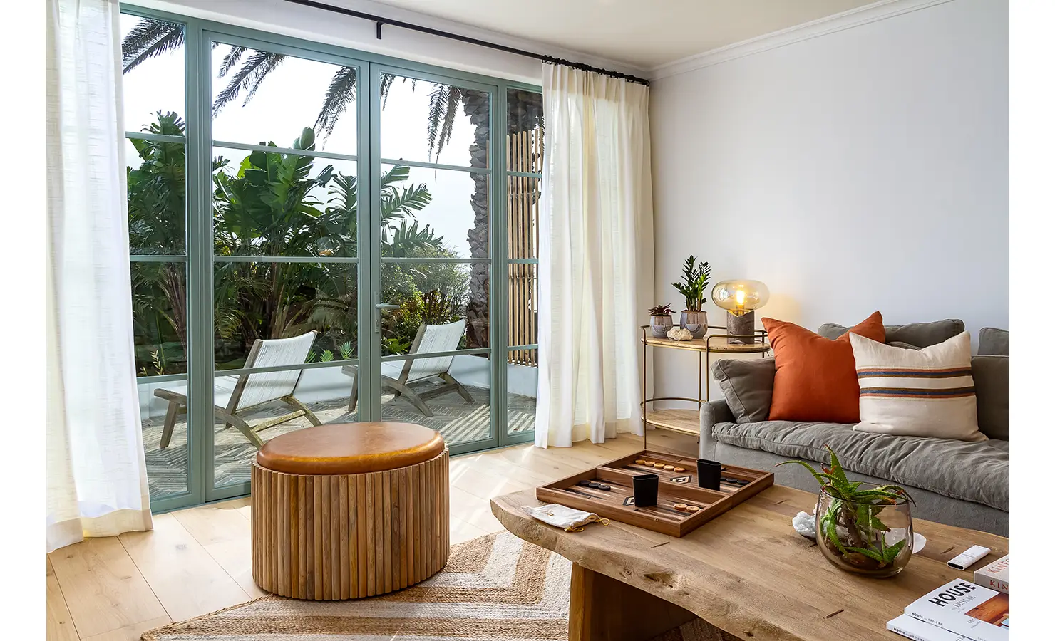 Lounge area with large wooden coffee table, natural rug and foot stool looking out glass doors. Solmar.