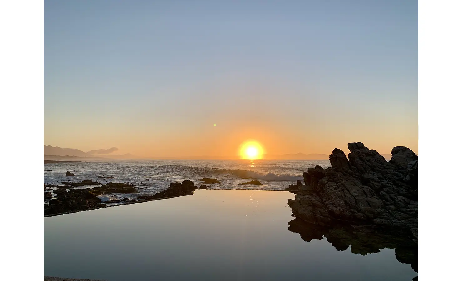 Still rock pool waters looking out to ocean and orange sunset. Cliffside. Hermanus.