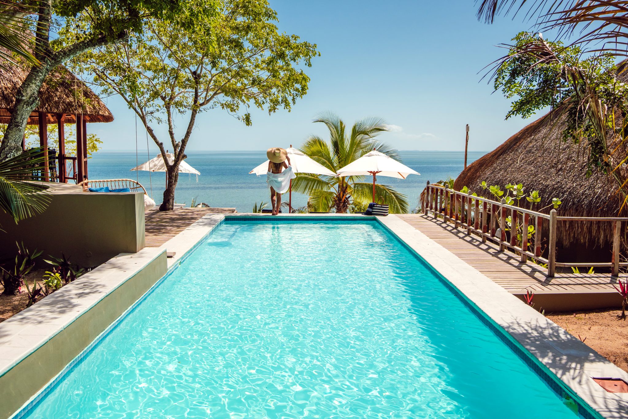 View from a large sparkling pool extending toward the ocean with palm trees and thatch lapas, lady in beachwear standing next to umbrellas at Mozawi Lodge Mozambique.