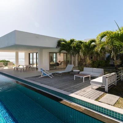 Villa De L'Ocean on the coast with ocean views, a lap pool, and an outdoor deck with loungers shaded by the palm trees.