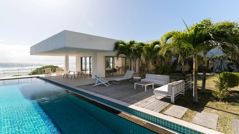 Villa De L'Ocean on the coast with ocean views, a lap pool, and an outdoor deck with loungers shaded by the palm trees.