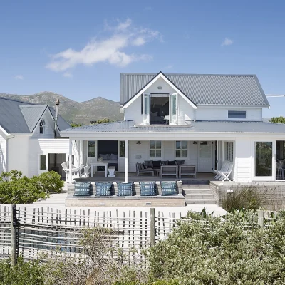 A classic white wooden two-story house with an extensive balau deck, large windows, and a fenced indigenous garden, set against a backdrop of mountains and greenery under a clear blue sky. Noordhoek Beach House.