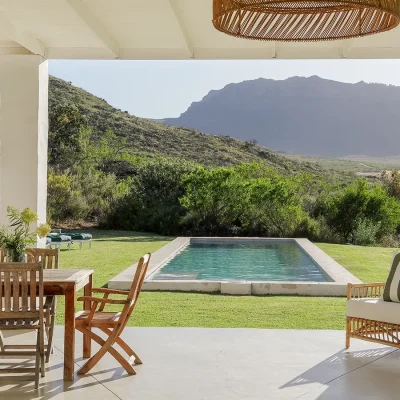 Buchubos Villa verandah with an outdoor wooden dining table and chairs overlooking the lap pool and green rolling lawn that is surrounded by the natural landscape and distant mountain range.