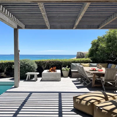 Oasis on Glen with an expansive wooden deck protected by a wooden pergola, a splash pool and a sunken fire pit with surrounding seating areas and panoramic views of a blue hued ocean meeting a crystal blue sky.