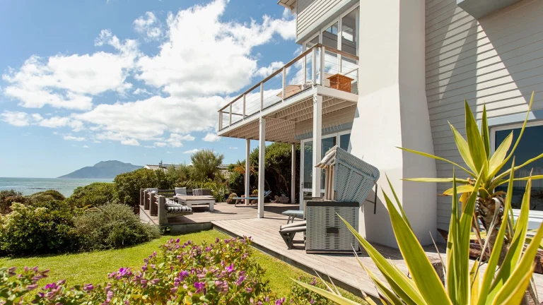 Pebbles, a beige clapboard home on the coast of Kommetjie with a wrap around verandah and green lawn.