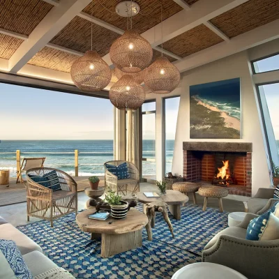 Lekkerwater beach lodge lounge matching the ocean front hue with a beige sofa and armchairs dressed in blue and white scatter pillows, a wooden coffee table and a blue and white floor rug facing the large indoor fireplace and surrounded by large stack back doors that reveals the ocean.