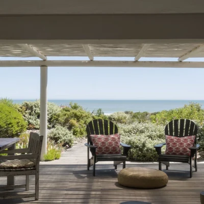 Wooden deck with wooden dining table and arm chairs shaded by a white bamboo pergola that opens onto a stretch of coastal vegetation and ocean views.