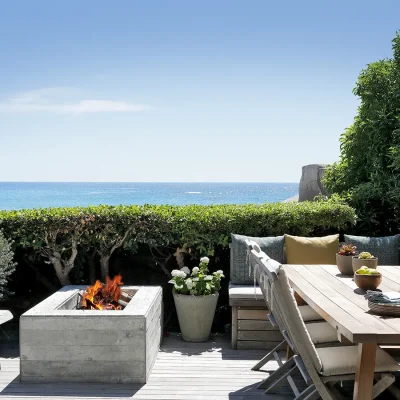 Outdoor fire pit with a wooden dining table and chairs shaded by a milkwood tree, and looking onto the crystal blue ocean. Oasis on Glen.