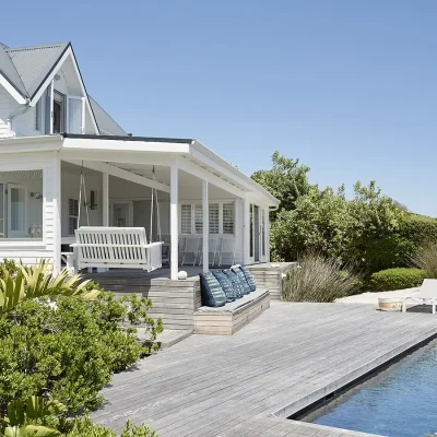 Noordhoek Beach House with white cladded walls, a porch swing overlooking a wooden deck and a swimming pool, surrounded by indigenous plants and patio chairs under a clear blue sky.