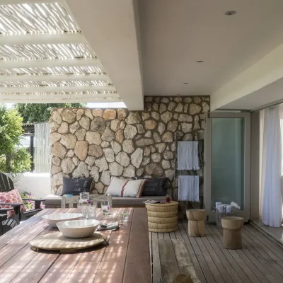 Large stack-back doors opening Beach House Zonnestraal lounge to a wooden deck furnished with a built-in corner sofa and wooden armchairs and a long dining table protected by a white bamboo pergola and corner stone wall.