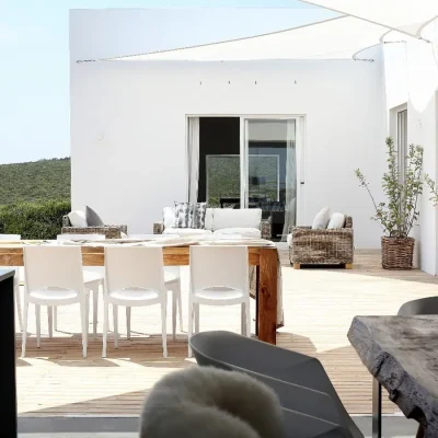 Protected wooden deck with white washed walls and furnished with outdoor wooden dining table and woven sofas with soft, white pillows. Feng shui.