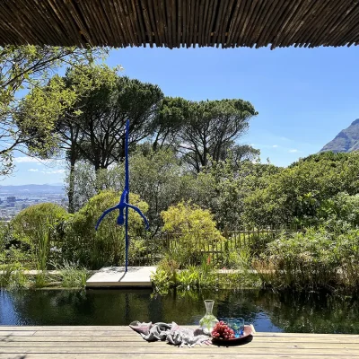 Wooden deck protected by a bamboo pergola overlooking a natural swimming pool with a statue and panoramic views of a luscious garden and distant city and Table Mountain. Villa Toscana.