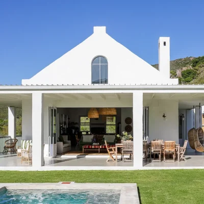 Buchubos Villa wrap around verandah with an outdoor wooden dining table, wicker sofa's and hanging wicker chairs with an outdoor braai, and a white finish.